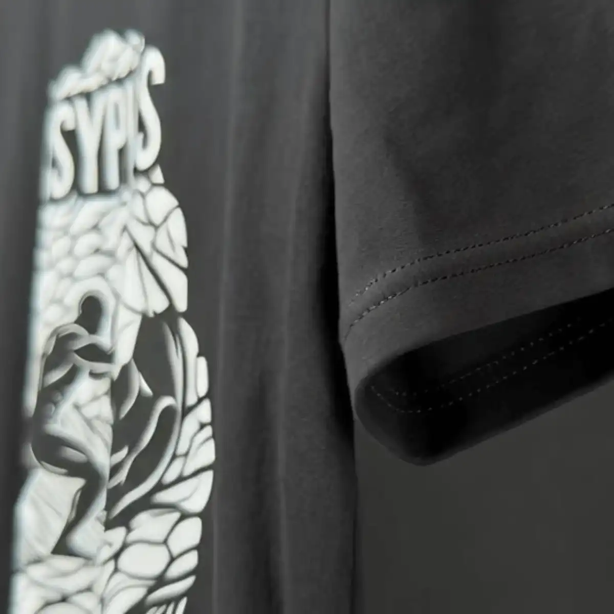 Camiseta Sísifo – Filosofia | Silk & Luck 3 Macro fotografia focada na bainha e costura da manga de uma camiseta preta, revelando parte da estampa branca geométrica ao fundo.